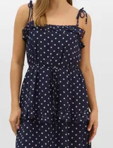Vestido largo azul marino con estampado de lunares blancos, confeccionado en gasa fluida. Cuenta con tirantes finos ajustables con lazos en los hombros, escote con volados, corte en la cintura con lazo ajustable, y falda larga con múltiples capas y volados horizontales.