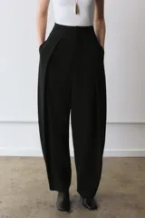 Pantalón negro de tiro alto, con pliegues en el delantero y bolsillos laterales ocultos. Cierre frontal con cremallera y ganchos metálicos.