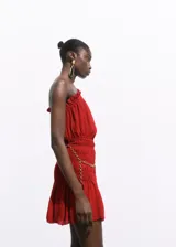 Vestido corto de color rojo, con diseño asimétrico de un solo hombro y detalle de volados en el escote. Presenta una sección central fruncida que se ajusta a la cintura y falda con vuelo. Incluye un cinturón decorativo de cadena metálica desmontable.