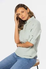Camisa blanca de manga larga con cuello y tapeta de medio botón. Presenta dos bolsillos frontales con tapa y botones metálicos. Las mangas tienen presillas para sujetar el puño enrollado.