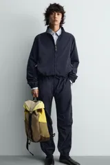 Mochila confeccionada en tejido técnico con bloques de color beige, amarillo y verde, y detalles en piel. Cuenta con un compartimento principal con cierre de solapa, hebilla y cordón ajustable, bolsillos laterales planos y correas de hombro regulables con hebilla de liberación rápida.