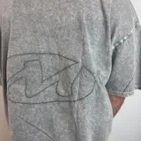 Remera de corte boxy fit con efecto lavado tipo snow wash, color gris. Presenta un diseño lineal bordado en la espalda con forma ovalada.