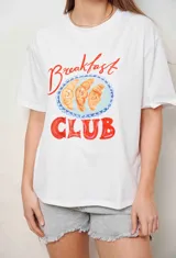 Remera blanca con diseño estampado de medialunas y texto 'Breakfast Club', con detalles en piedritas bordadas y mangas cortas.