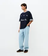 Remera de tejido texturizado color azul marino, con cuello redondo y mangas cortas con puños a rayas blancas. Presenta bordados frontales con la inscripción 'Future Co.', el número '98', un logo ovalado y estrellas.