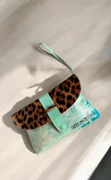 Sobre de mano de cuero turquesa con solapa de animal print de leopardo y tira central texturizada. Incluye detalles de arte pintado a mano, inscripción de la marca y correa de muñeca.