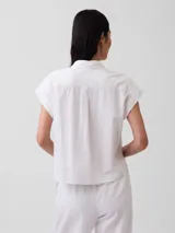 Camisa de lino blanca de manga corta, con cuello camisero, cierre frontal de botones y corte corto.