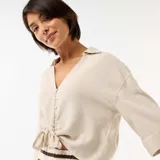 Camisa corta oversize color beige, con mangas largas, cuello camisero, cierre frontal de botones y detalle de nudo en el frente.