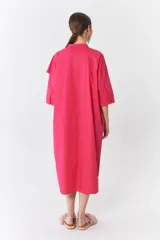 Vestido camisero fucsia de poplín de algodón con manga 3/4, cuello mao, cierre frontal con botones y bolsillos laterales. Presenta un volado diagonal en el delantero.