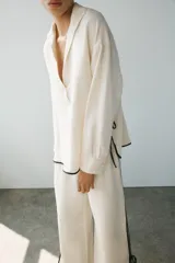 Camisa de lino color crudo con cuello solapa, manga larga y bajo asimétrico. Presenta detalles de ribetes negros a contraste y aberturas laterales con tiras para ajustar.
