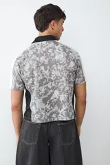 Camiseta de estilo fútbol con estampado de camuflaje gris, cuello tipo polo negro y mangas blancas. Presenta gráficos frontales con la palabra LEGACY en amarillo y detalles de escudos deportivos.