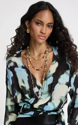 Blusa de tejido fluido con estampado abstracto en tonos azules, negros y beige. Presenta un diseño cruzado con escote en V, mangas 3/4 anchas y un lazo lateral ajustable que define la silueta.