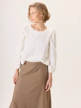 Camisa blanca de algodón con mangas largas abullonadas, detalle de volados en los hombros y puños elásticos. Presenta un diseño con cuello redondo y corte holgado.