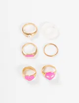 Set de anillos dorados con apliques de plástico rosa y blanco. Incluye un anillo con forma de corazón sonriente, un anillo con forma de alien, un anillo con aplique rectangular, un anillo con perla y dos anillos lisos.