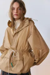 Cazadora tipo anorak color beige, con capucha ajustable, manga larga y bolsillo frontal de plastrón con solapa. Cuenta con cordones elásticos ajustables en la cintura y el bajo, y cierre frontal con botones a presión.