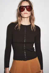 Chaqueta ajustada de punto acanalado color marrón oscuro, con cuello redondo y manga larga. Presenta un cierre frontal completo con botones pequeños y blancos. Los bordes del ruedo y los puños tienen un acabado festoneado.
