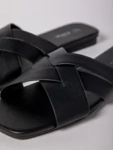 Sandalia baja negra de cuero sintético, con tiras anchas cruzadas en el empeine y suela plana.