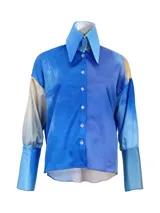 Conjunto de tres piezas inspirado en la vestimenta tradicional uruguaya, compuesto por camisa, pantalón tipo chiripá y accesorio, confeccionado en tela satinada con estampado artístico sublimado en tonos azules y amarillos, con terminaciones de flecos en color marrón.