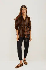 Saco tipo camisa de algodón con rayas verticales marrones y negras. Presenta bordados de cuentas en tonos tierra en el hombro izquierdo y se ajusta a la cintura con un lazo de viscosa estampado en tonos naranjas y negros.