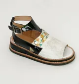 Sandalia de cuero Carolina De Cunto modelo Leyla, con semi plataforma y diseño combinado en charol negro, cuero blanco y una franja con detalles pintados a mano en tonos dorados y multicolores. Cuenta con tira ajustable en el talón con hebilla metálica.