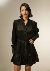 Vestido corto de satén negro, con cuello mao y detalle de volados en la botonadura frontal. Posee mangas largas con puños ajustados y un cordón fino en la cintura para entallar, terminado con pequeñas cuentas doradas.