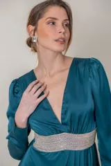Vestido largo de fiesta en satén de seda color petróleo, con escote en V profundo, mangas largas y falda cruzada con abertura. Presenta aplique de strass en la cintura.