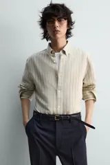 Camisa de algodón de corte relajado, con cuello solapa abotonado (button-down) y manga larga terminada en puño con botón. Presenta un patrón de rayas verticales finas en tonos beige claro y marrón/verde oliva.