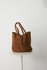 Bolso tipo tote de cuero gamuzado color tostado, con diseño de doble asa y cierre mediante cordón ajustable en la parte superior.
