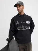 Buzo de manga larga color negro con cuello tipo polo y detalles de líneas blancas en cuello y puños. Presenta un diseño gráfico estampado en el frente con la inscripción 'CHALÉT CLUB' y detalles deportivos. Confeccionado en tejido suave con ajuste holgado.