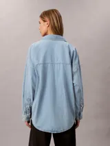 Camisa de denim de corte holgado, con cuello en punta, mangas largas y cierre frontal con botones. Presenta un diseño clásico con acabado lavado y monograma bordado en el pecho.