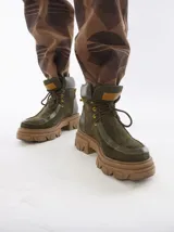 Borcegos de caña baja con diseño robusto, confeccionados en una combinación de gamuza y cuero sintético en tonos verde oliva. Presentan suela track de goma dentada en color suela, cordones al tono con ojalillos amarillos y cuello acolchado para mayor confort.