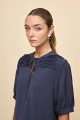 Blusa de manga corta color negro, con calce relajado, escote redondo con abertura y lazo para anudar. Presenta detalles de tablas horizontales en la parte superior que aportan textura al diseño.