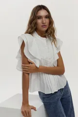 Blusa blanca sin mangas, con volados amplios en los hombros, bordado a tono en el pecho, detalles de tablones a lo largo del cuerpo y cuello elevado con volados.