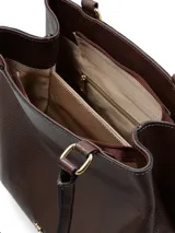 Cartera de hombro color chocolate con textura granulada, asas fijas y detalle decorativo de nudo con cordones en tono rosado en uno de los laterales. Incluye logo metálico en la parte inferior frontal.