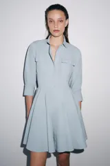 Vestido corto tipo camisero de algodón, con cuello solapa, mangas tres cuartos y cierre frontal con botones. Presenta bolsillos de plastrón con solapa en el pecho y falda con bajo plisado.