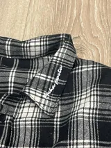 Camisa de manga larga con corte boxy fit, confeccionada en algodón a cuadros en tonos negro y gris. Presenta un bolsillo frontal en el pecho y el texto 'posta.' estampado en color blanco en la parte delantera.