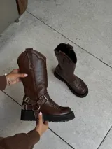 Bota de caña alta confeccionada en cuero vacuno engrasado color chocolate con efecto desgastado. Presenta bordados artesanales en la caña y el empeine, y cuenta con un sistema de correas con hebillas metálicas desmontables. Posee una suela track de goma negra de estilo robusto.