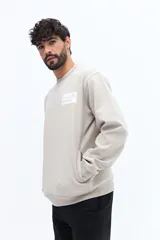 Buzo de algodón y poliéster color verde musgo, con cuello redondo y puños acanalados. Presenta un parche estampado rectangular con texto y un logo circular en el pecho.