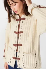 Cardigan de punto color crudo con textura acanalada, cierre frontal con botones tipo toggle en color marrón y bolsillos frontales. Presenta detalles de costuras y terminaciones en color marrón.