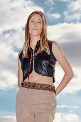 Chaleco corto negro de cuero ovino, con cuello mao y cierre frontal mediante tientos de cuero marrón que pasan por ojalillos metálicos.