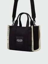 Cartera tipo tote bag de lona color beige, con diseño estructurado y asas superiores. Presenta el logo de la marca estampado en la parte frontal y detalles de cintas laterales con inscripciones.