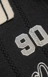 Sweater de punto negro con cuello polo en color crema, manga larga y diseño oversize. Presenta bordados en el frente con la inscripción 'Ravers', el número '90' y letras cursivas, con terminaciones en punto rib en puños y cuello.