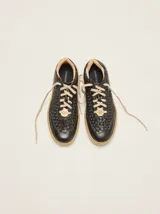 Championes urbanos de cuero negro con diseño trenzado en los laterales y la puntera, cordones en tono tostado y suela de goma. Cuentan con un detalle de aplique metálico dorado en los cordones.