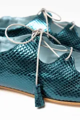 Zapato tipo blucher con diseño híbrido de bailarina, confeccionado en piel azul con textura de reptil. Presenta cierre con cordones y borlas de piel, con una banda elástica en la parte trasera para un ajuste óptimo. Cuenta con suela de cuero y acabado artesanal.