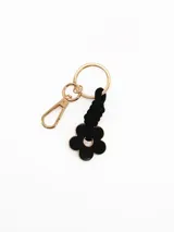Llavero con argolla y mosquetón metálicos dorados, unido a un dije en forma de flor negra con borde dorado mediante un cordón negro trenzado.