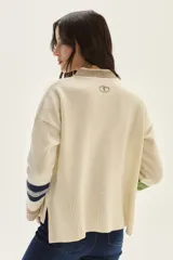 Sweater tejido de lambswool color beige, con cuello redondo y puños acampanados con rayas de colores (marrón, mostaza, bordó y beige). Incluye un detalle de temporada hecho a mano en la espalda.