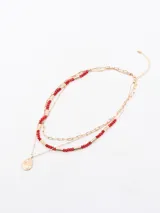 Collar multicapa de color dorado, compuesto por una cadena de eslabones finos y una cadena con cuentas rojas y cilindros dorados, con un dije en forma de gota texturizada.