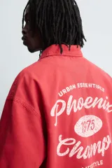 Camisa de algodón con estructura canvas, color rojo lavado, de corte relajado (relaxed fit) y manga corta. Posee cuello solapa, dos bolsillos de plastrón en el pecho y cierre frontal de botonadura. Presenta bordado y estampado de texto a contraste en el delantero.