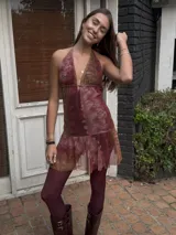 Vestido corto negro con escote en V profundo, estilo halter con tiras para atar al cuello. Presenta un diseño ajustado en el busto con tiras ajustables bajo el mismo que se cruzan y anudan en la espalda, y falda con volado en el ruedo con detalle de encaje.