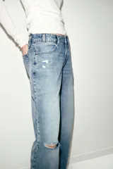 Jeans de tiro bajo, corte recto y pierna ancha, color celeste claro desgastado. Presenta roturas visibles en la zona de la rodilla y pequeños desgastes en los muslos y bolsillos.