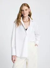 Camisa blanca de corte recto y estilo oversized, con cuello clásico, mangas largas con puños anchos, bolsillo delantero, cierre frontal con botones ocultos y bajo ligeramente redondeado.